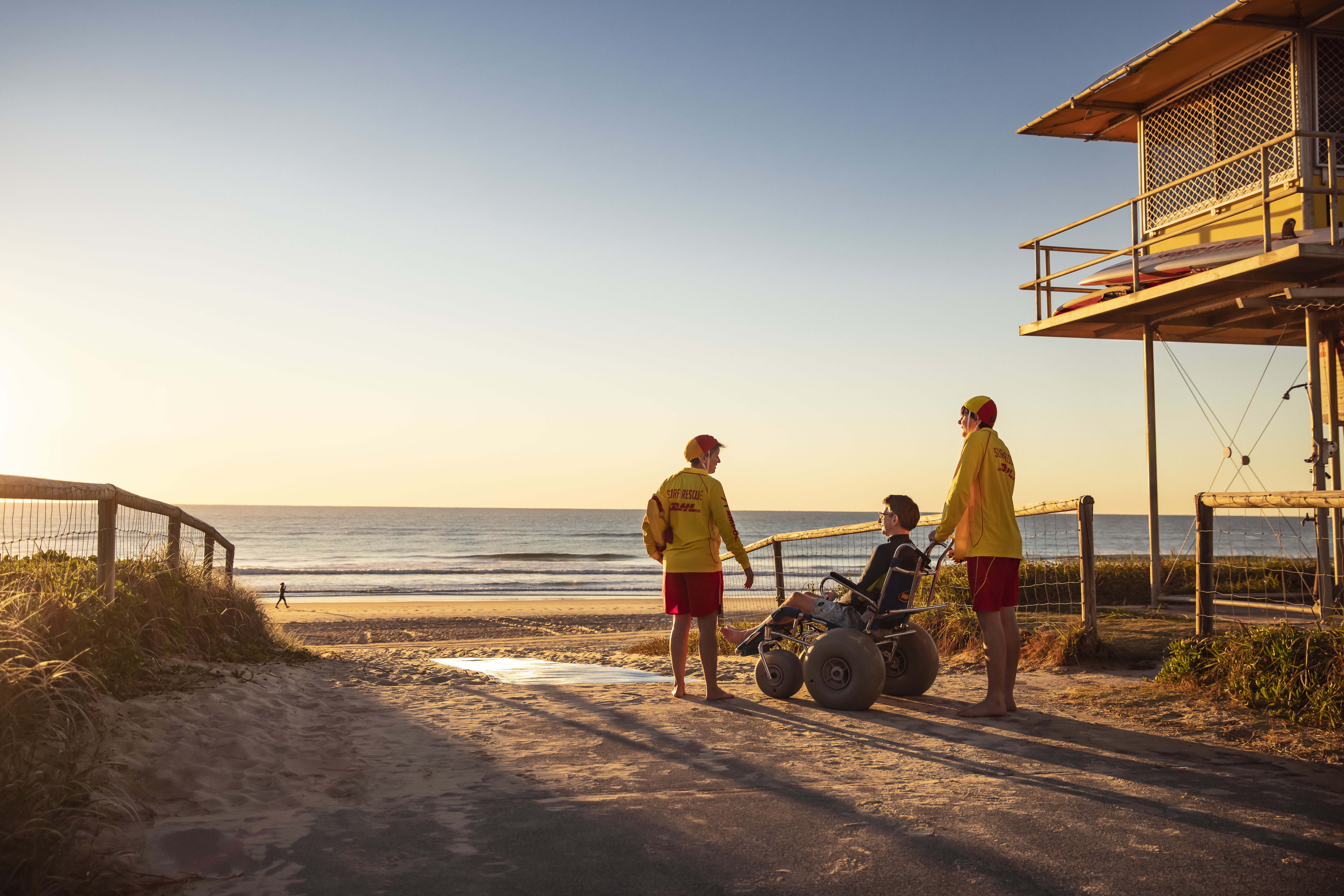 Sunset beach theme with two lifesavers chatting with a man in a beach wheelchair next to the life-saving tower