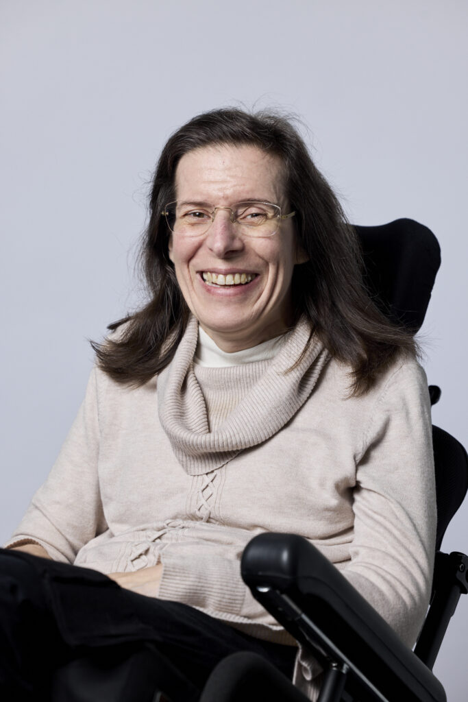 Yasmine Gray GetAboutAble founder and AITCAP creator dressed in a beige pullover and smiling, seating in her powerwheelchair.